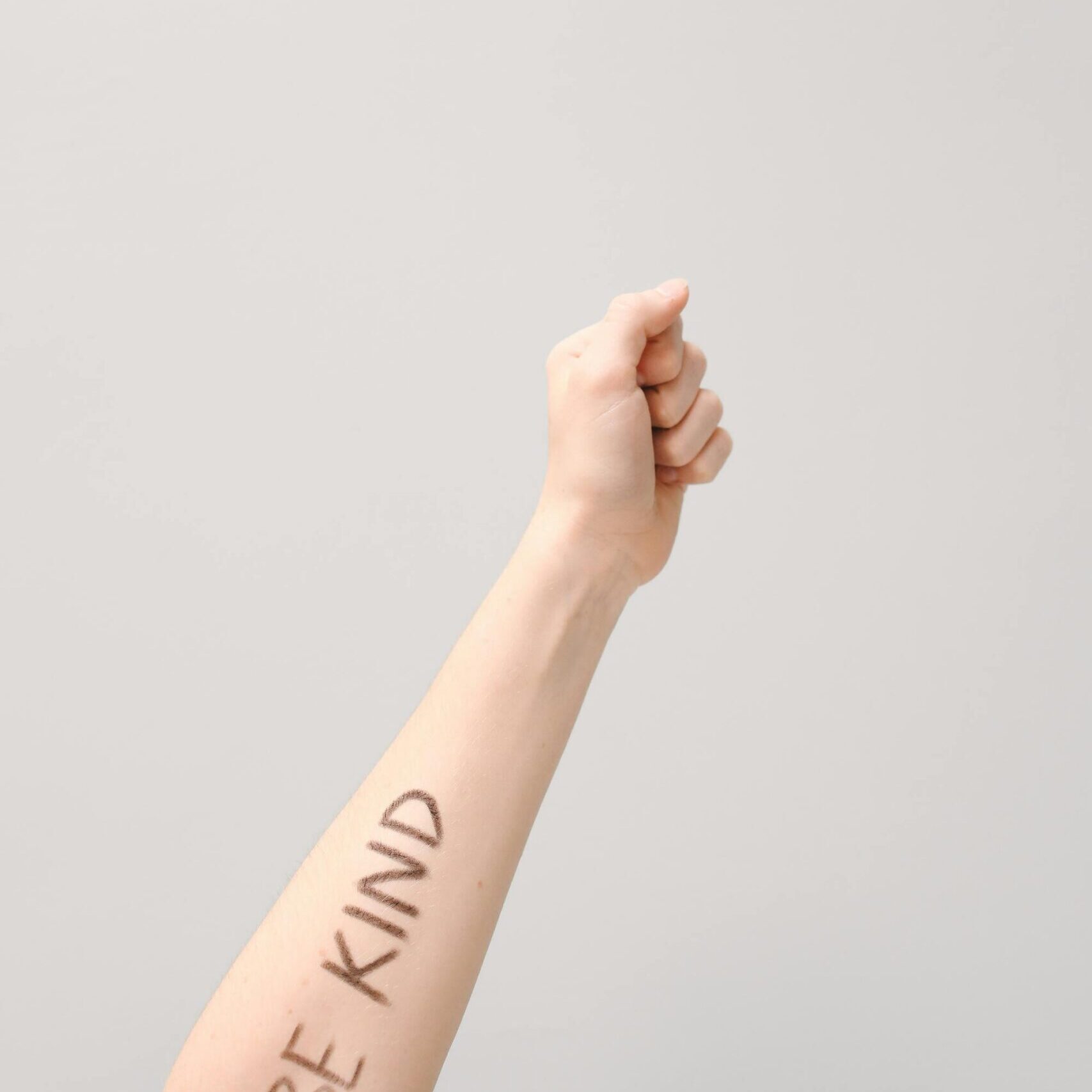 Raised arm with 'Be Kind' message written in minimalist studio setting. Emphasizes kindness and positivity.